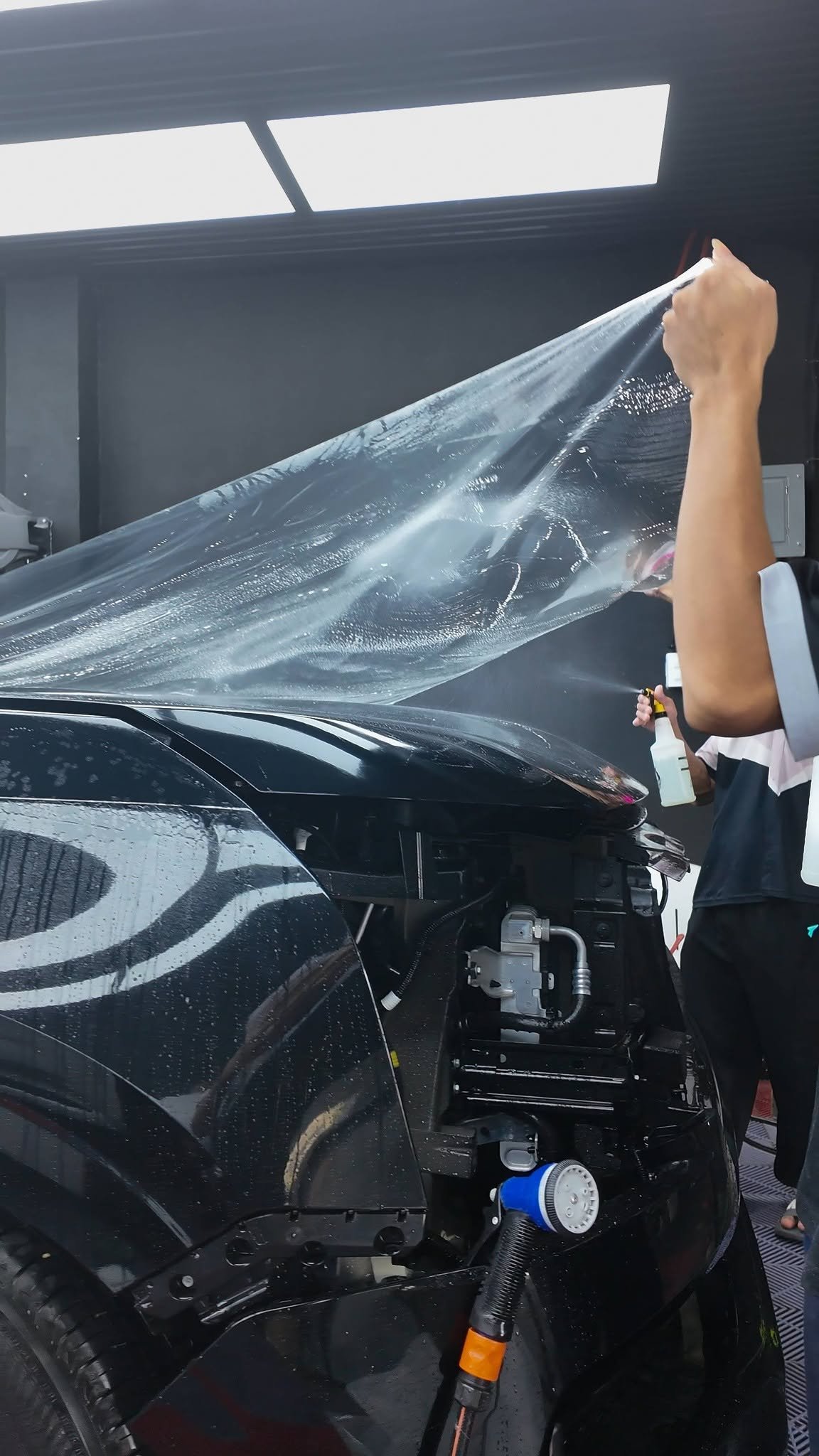 A technician floating a sheet of clear paint protection film over the wet hood of a black car