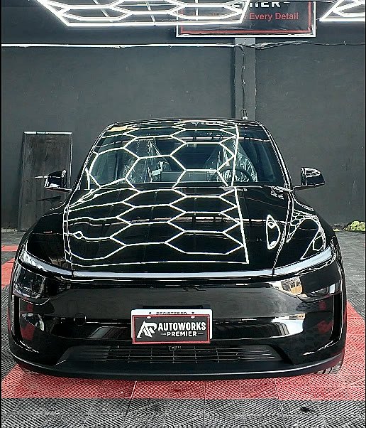 A black Tesla Model Y seen head-on under the hexagon lights, the light frames reflecting evenly across a full body paint protection film finish
